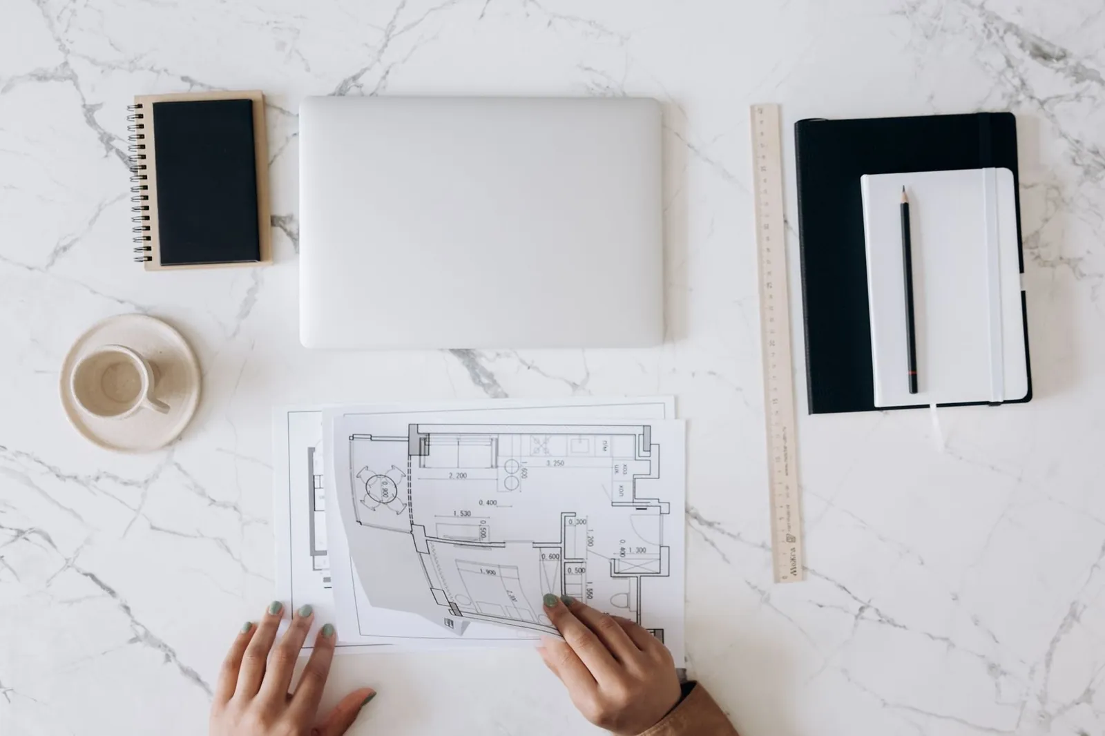 A marble desk from above with a laptop, notebooks, a ruler and hands on a floor plan